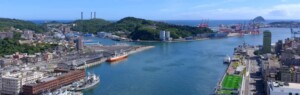 Aerial view of a coastal port city with cranes, quays, and several ships along a calm blue harbor.